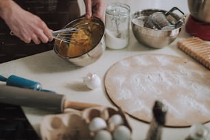 Kitchen table with flour sugar rolling pins and bowls. Person whisking eggs