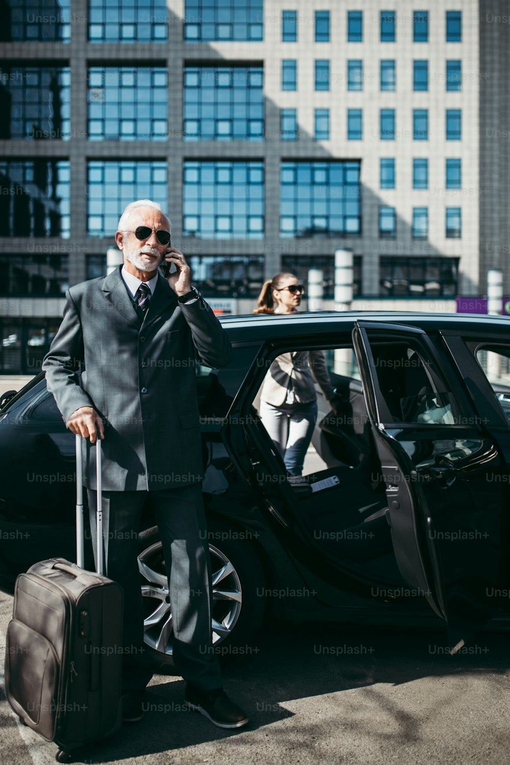Gut aussehender älterer Geschäftsmann und seine junge Kollegin oder Kollegin stehen am Auto. Er telefoniert mit dem Smartphone, hält einen Koffer und bereitet sich auf eine Geschäftsreise vor. Transport im Unternehmenskonzept.