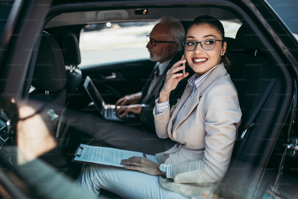 高級車の後部座席に座っている見栄えの良い年配のビジネスマンと彼の若い女性の同僚または同僚。彼らは話し、笑顔で、ラップトップやスマートフォンを使用しています。企業事業のコンセプトにおける輸送。