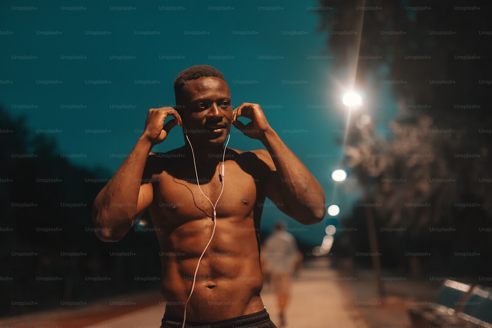 handsome smiling african shirtless male runner putting earphones in ears while standing on the racetrack at night. Nothing tastes as good as fit feels.