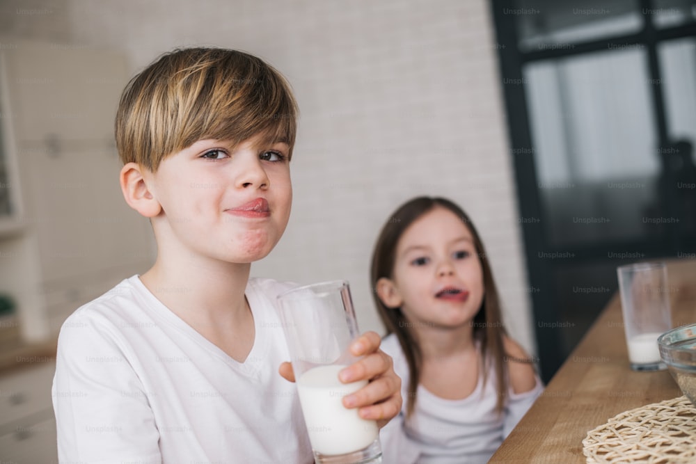 Cropped photo of little sister and brother holding glasses of milk while sitting at table indoors. Visiting grandmother concept