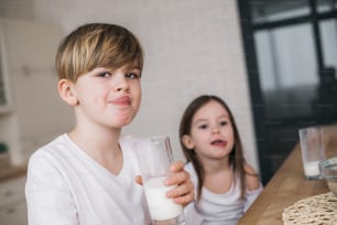 Cropped photo of little sister and brother holding glasses of milk while sitting at table indoors. Visiting grandmother concept