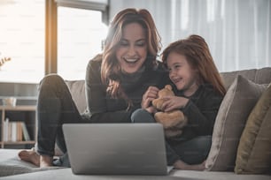 Hilarious. Smiling red-haired woman sitting on couch at home with her little child. They are looking at something on laptop and having fun
