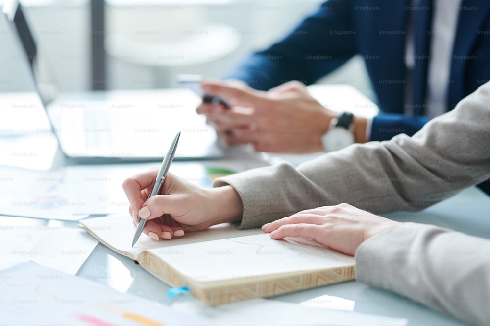 Hands of young elegant female economist making organization notes in notebook while sitting by desk in working environment