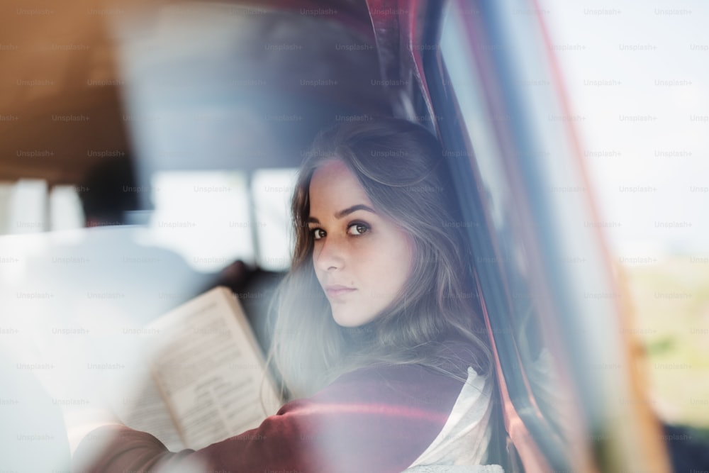 Un grupo de jóvenes amigos con una minivan retro en un viaje por carretera por el campo, una chica leyendo un libro. Disparo a través del vidrio.