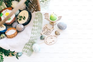 Stylish Easter eggs, easter bread cake, ham, beets, sausage, butter, green branches in wicker basket on white wooden background with easter candle and decorations. Happy Easter