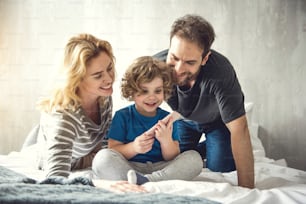 Doting parents are watching smiling son handling mobile phone. Parents and kid are sitting in light room. Little boy is showing interest to modern gadget