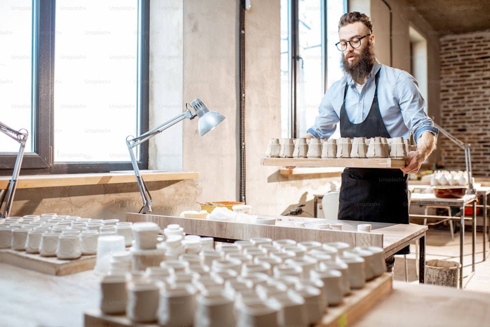 Handsome bearded man carrying tray with ceramic workpieces at the pottery shop