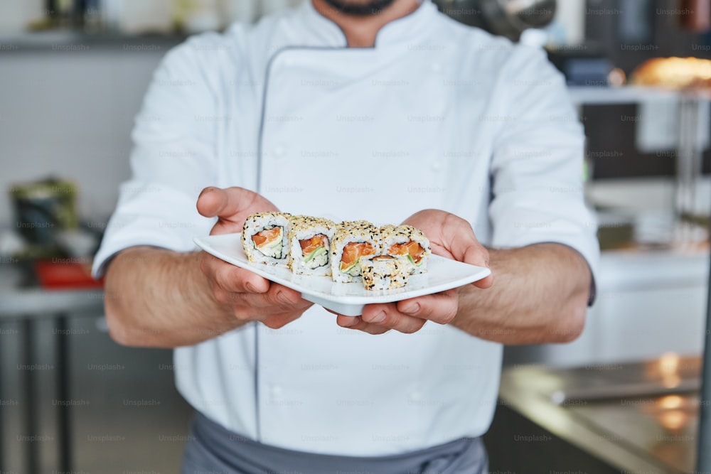 Chef japonês fez sushi no restaurante. Ele está de pé na cozinha comercial e segurando seu prato. Imagem recortada. Vista frontal
