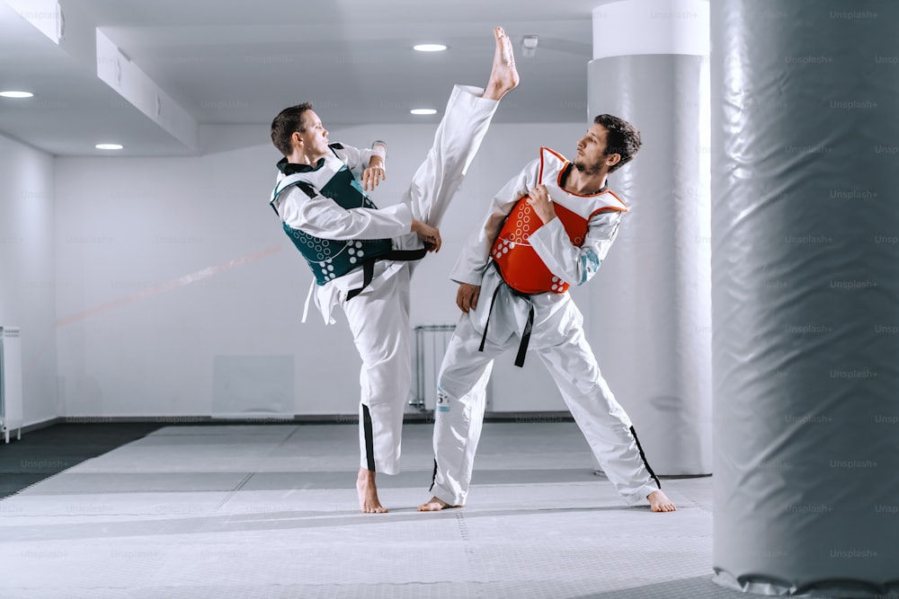 Dois homens esportivos caucasianos brigando em encaixes de tekwondo descalços.