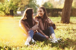 Jeunes jumelles brunes souriantes assises sur l’herbe avec les jambes croisées et légèrement pliées dans les genoux vêtues d’un manteau décontracté, bavardant, se regardant dans un parc ensoleillé d’automne sur fond flou.