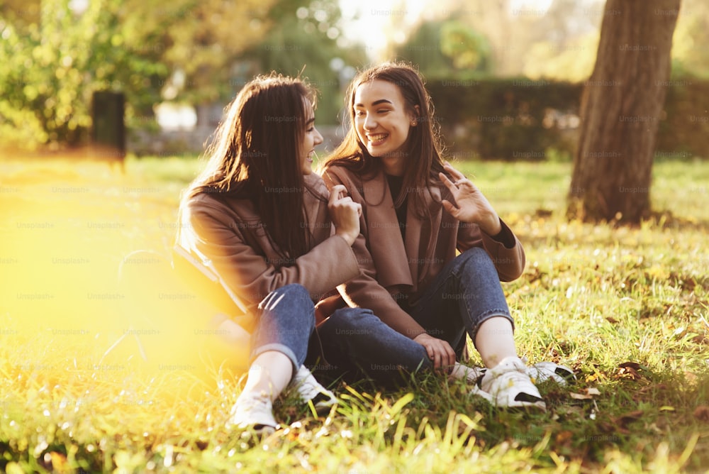 Young smiling brunette twin girls sitting on grass with legs crossed and slightly bent in knees wearing casual coat, chatting, looking at each other at autumn sunny park on blurry background.