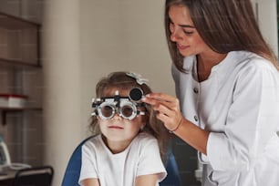 Der Arzt stimmt das Gerät ab. Kleines Mädchen mit Brille, das in der Klinik sitzt und ihre Augen testen lässt.