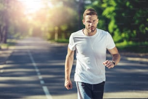 Healthy lifestyle. Portrait of young guy is running outdoors while listening to music through earphones. He is looking at camera confidently. Copy space in left side and sunshine in background