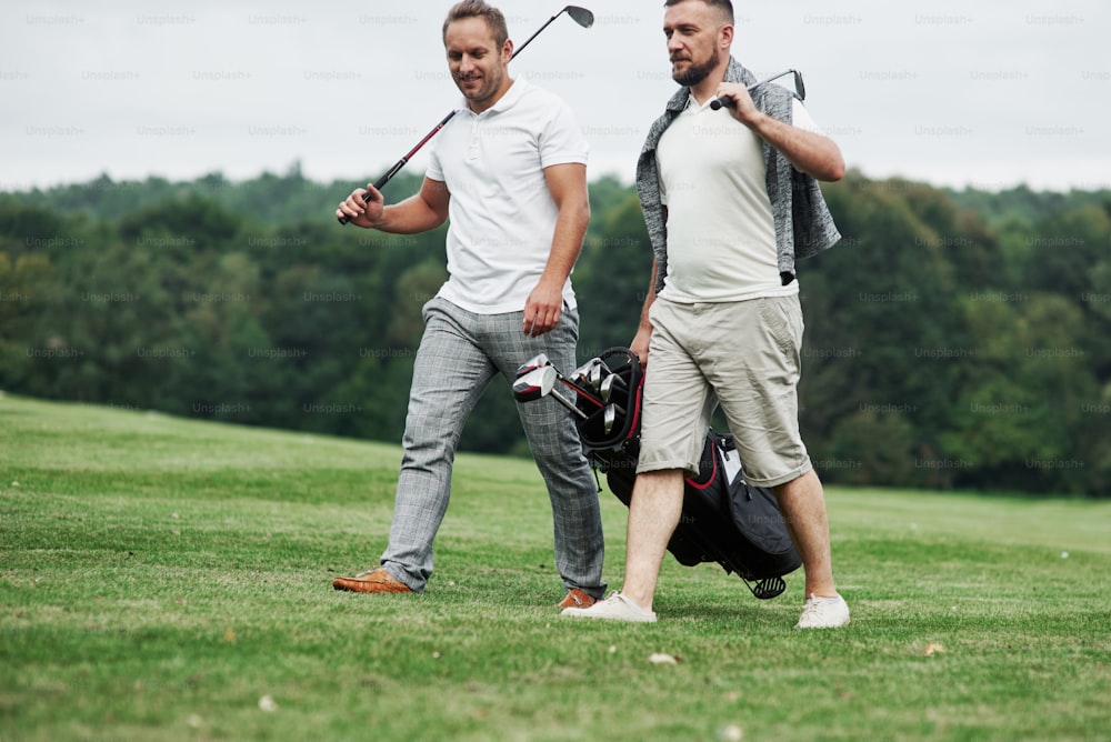 Two friends walking through the lawn with golf equipment and talking.