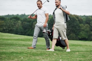 Two friends walking through the lawn with golf equipment and talking.