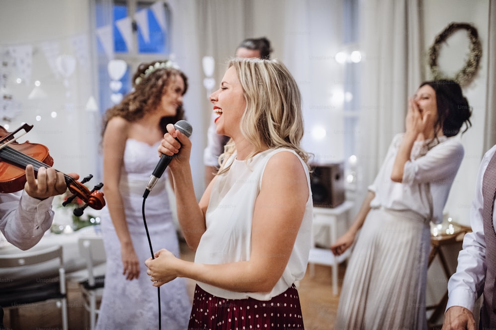 Uma jovem mulher cantando em uma recepção de casamento, noiva e convidados dançando ao fundo.