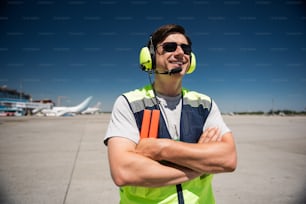 Enjoying beautiful sunny day. Waist up portrait of smiling man in headphones and sunglasses. Blue sky, runway and planes on background
