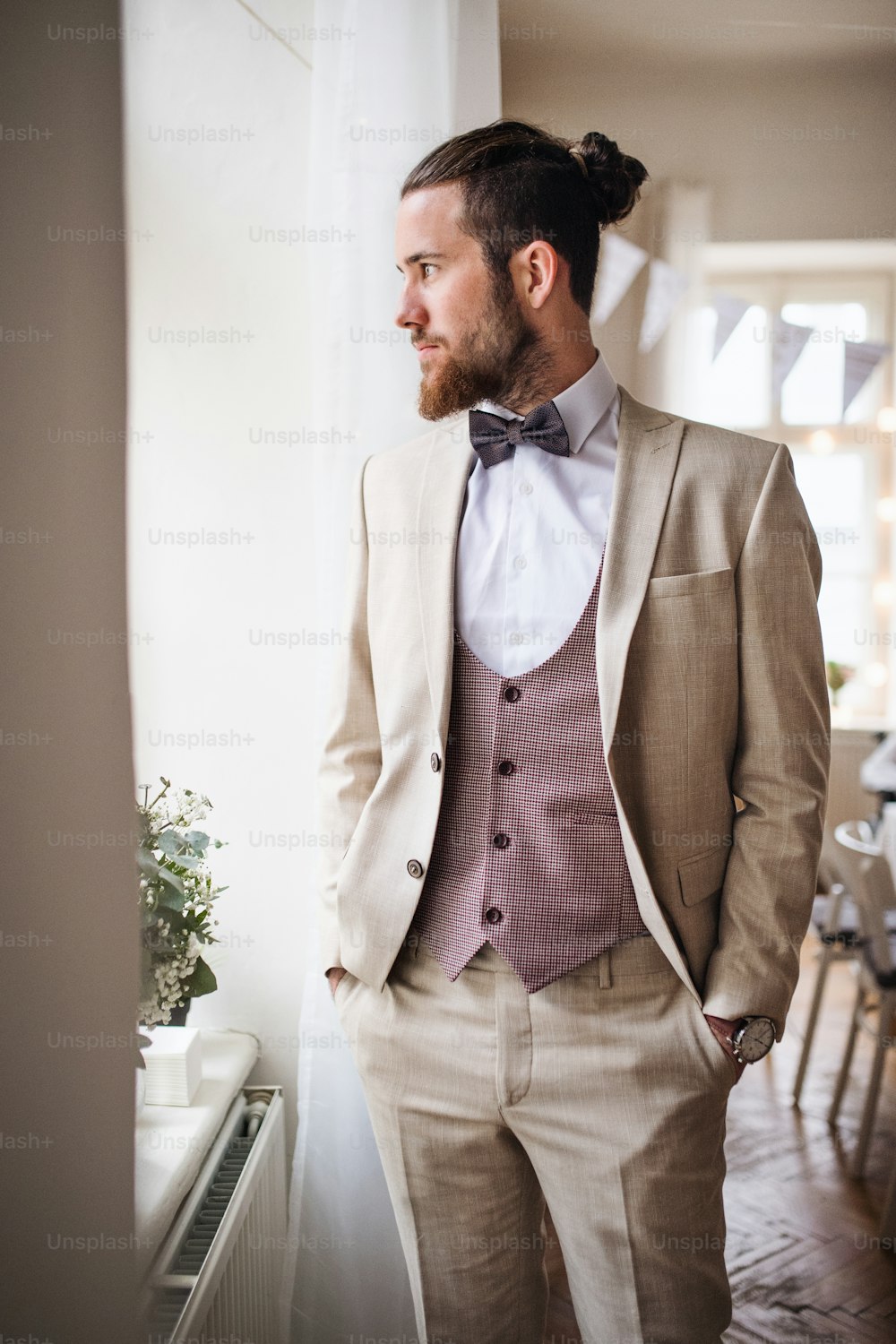 A handsome hipster young man with formal suit standing by a window on an indoor party, looking out.