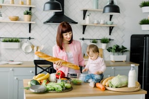 Mothers Day, healthy eating, cooking and family concept - happy mother fresh baguette and little baby girl siting on the dinner table with fresh vegetables, fruits and pot at home kitchen