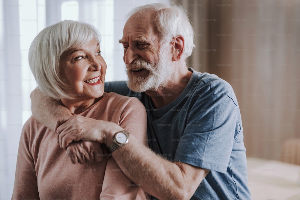 Love and tendance in any age. Close up portrait of happy smiling gray haired male embracing his beloved female partner from back while being at home