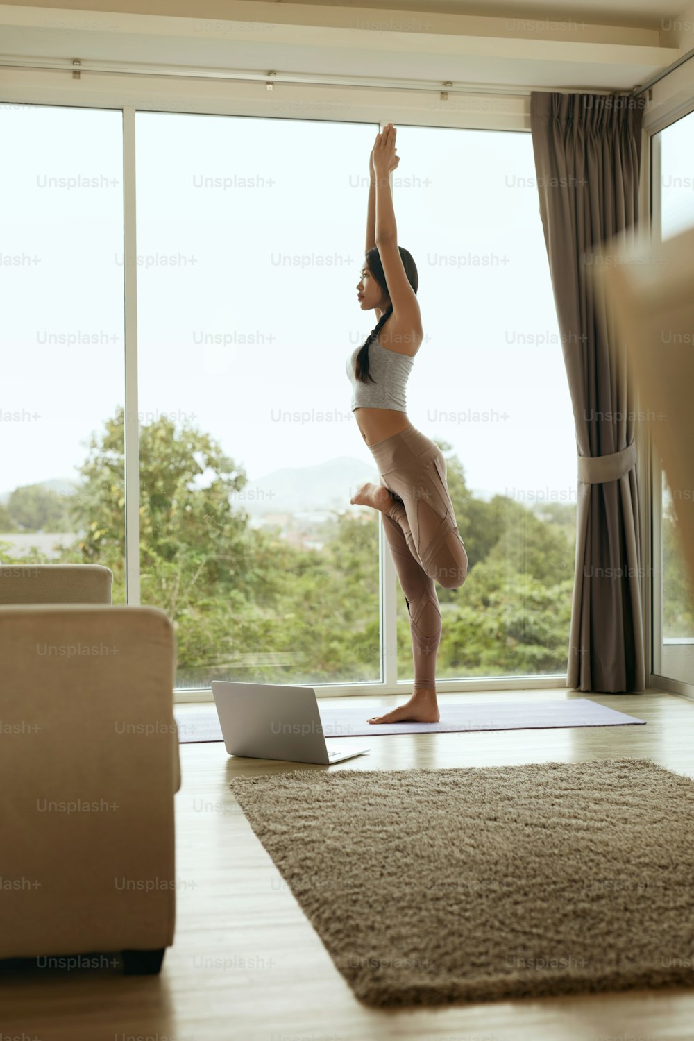 Girl practices yoga in tree pose, exercising at home in morning. Beautiful asian woman meditating in living room near laptop computer on ground