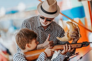 Grandfather and grandson having fun and spending good quality time together in amusement park. Kid shooting with air gun while grandpa helps him to win the prize.