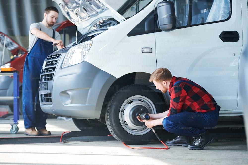 Two young professional car repair technicians serving one of automobiles in hangar or workshop