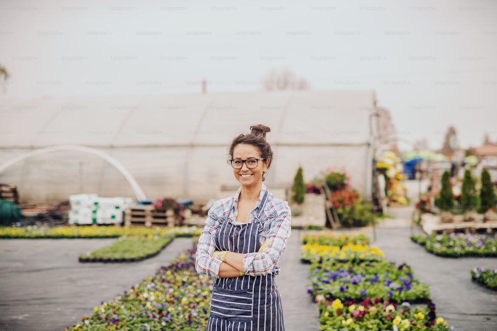 Charming Caucasian florist standing outdoors with arms crossed. In background greenhouse and pots with flowers.
