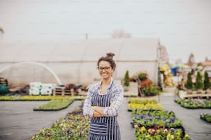Charming Caucasian florist standing outdoors with arms crossed. In background greenhouse and pots with flowers.