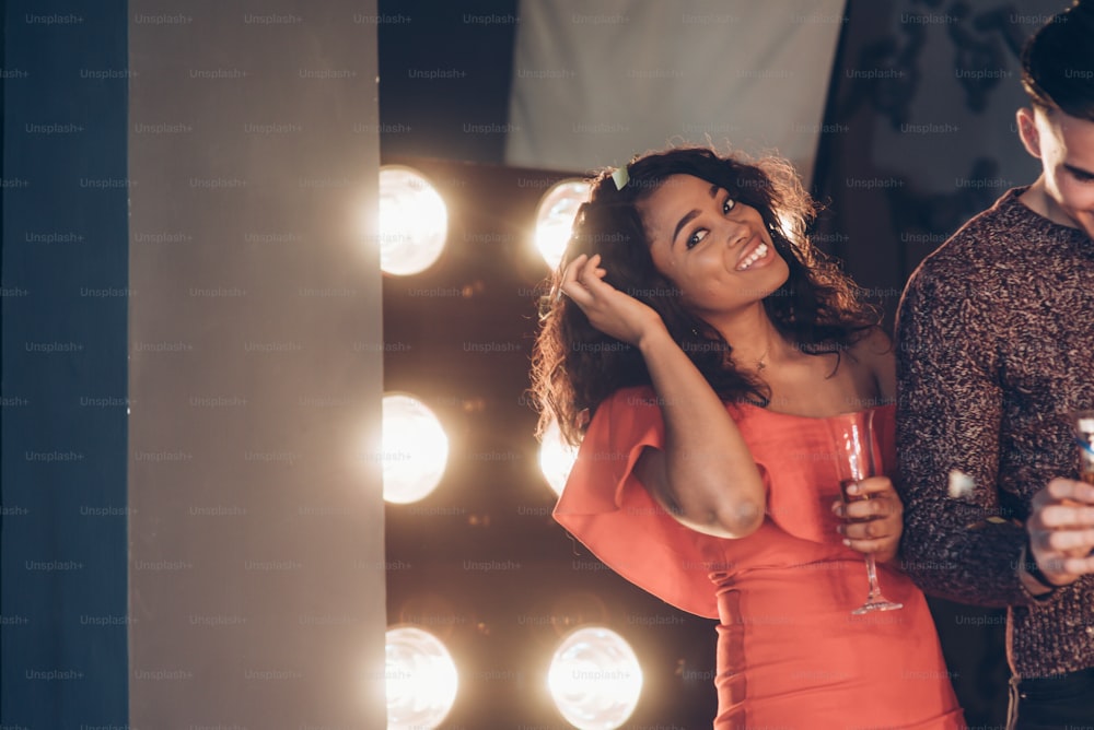 Hermosa joven afroamericana con vestido rojo mirando a la cámara y sonriendo mientras camina con un amigo y sostiene el vaso de bebida.