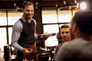 Young smiling waiter serving male guests and talking to them in coffee shop.