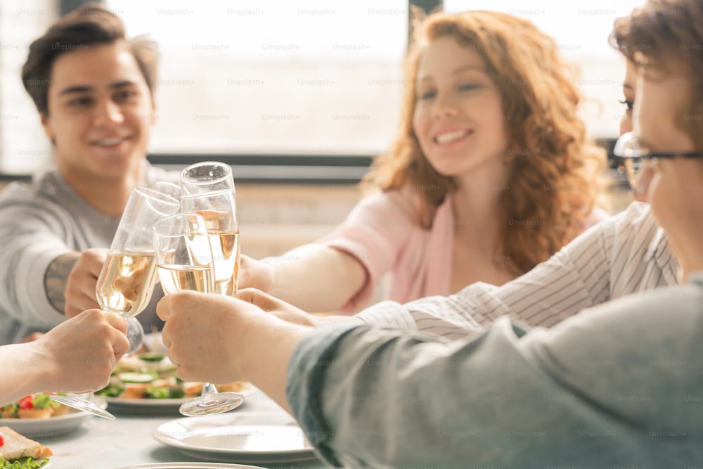 Group of friends clinking with flutes of champagne over homemade snacks on served table while celebrating life event