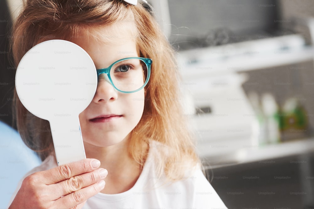 Try to read that word. Woman hands holds the occluder. Little girl checking his vision with new green glasses.