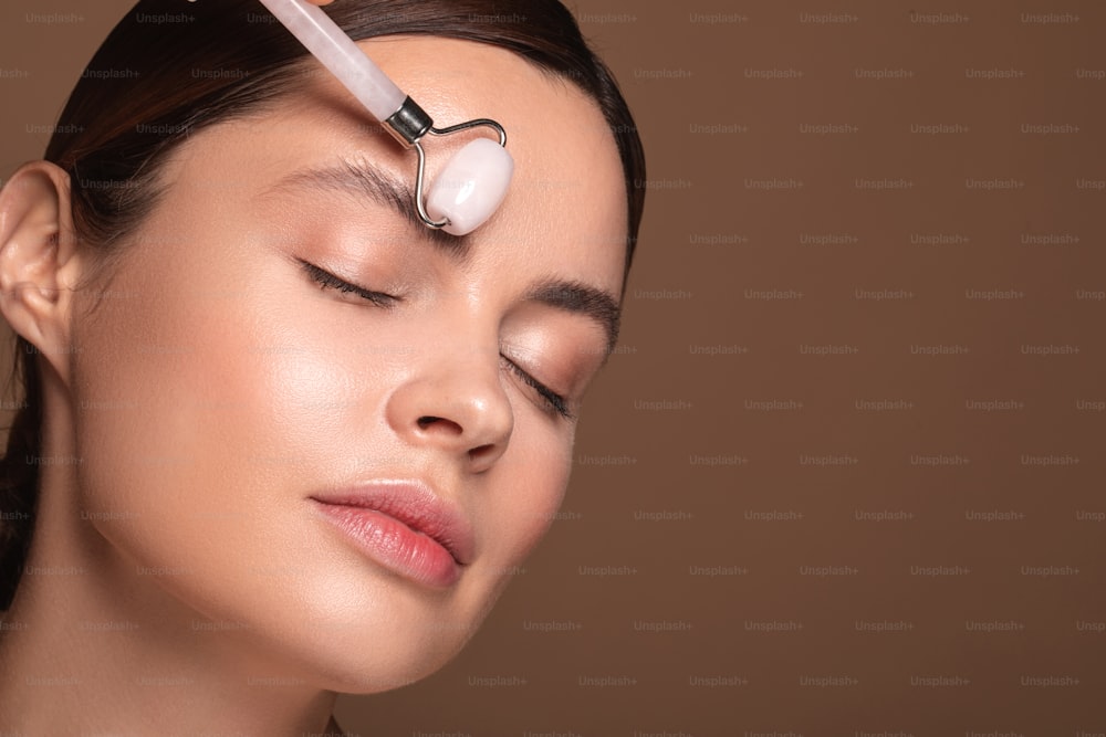 Relaxed lady isolated on the brown background having her eyes closed and massaging forehead with a face roller