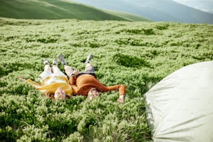 Lovely couple in bright sweaters lying on the green meadow near the campsite in the mountains