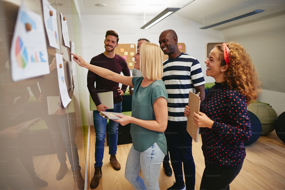 Diverse group of smiling colleagues looking at graphs and charts taped to a wall while having a meeting together in a modern office