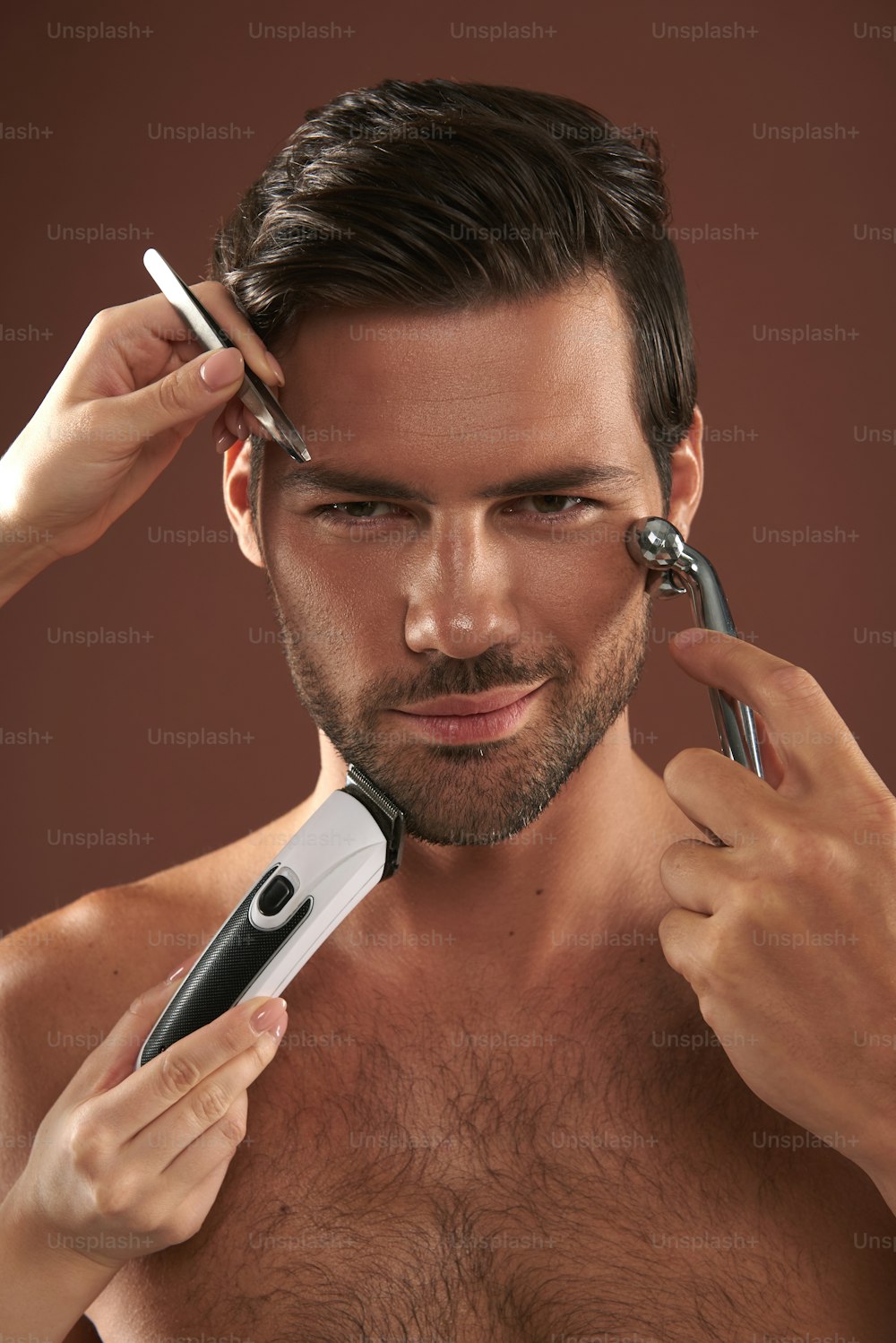 Retrato de primer plano de un macho sonriente que se afeita con una navaja de afeitar mientras alguien se depila las cejas y se afeita. Aislado en marrón