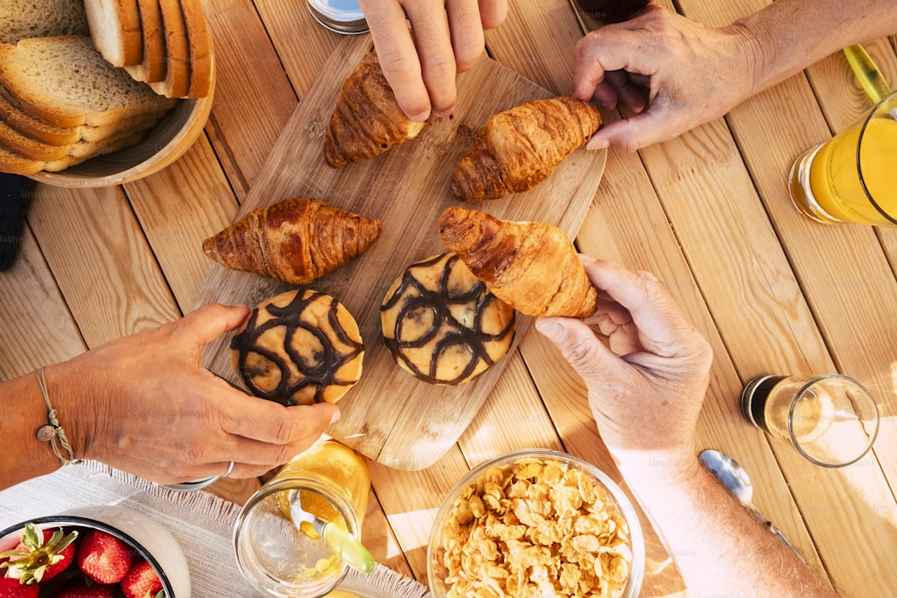 Groupe d’amis, membres de la famille, vue de dessus verticale, prenant des croissants et des aliments mélangés pour le petit-déjeuner, activité matinale - table en bois en arrière-plan et mains prenant pour manger