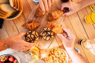 Group of friends family people viewed from vertical top view taking croissant and mixed food for breakfast morning activity - wooden table in background and hands taking to eat