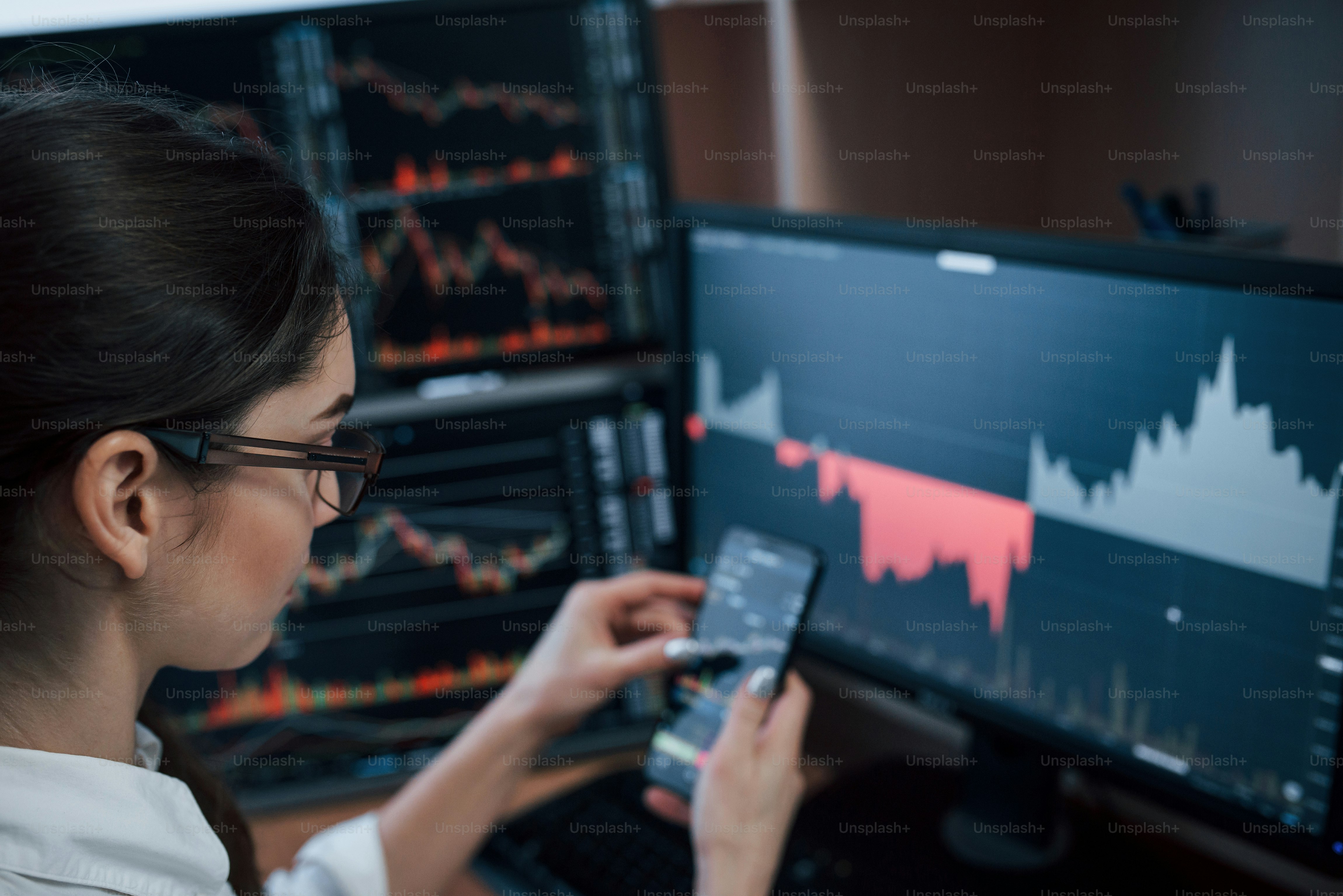 In eyeglasses. Woman working online in the office with multiple computer screens in index charts.