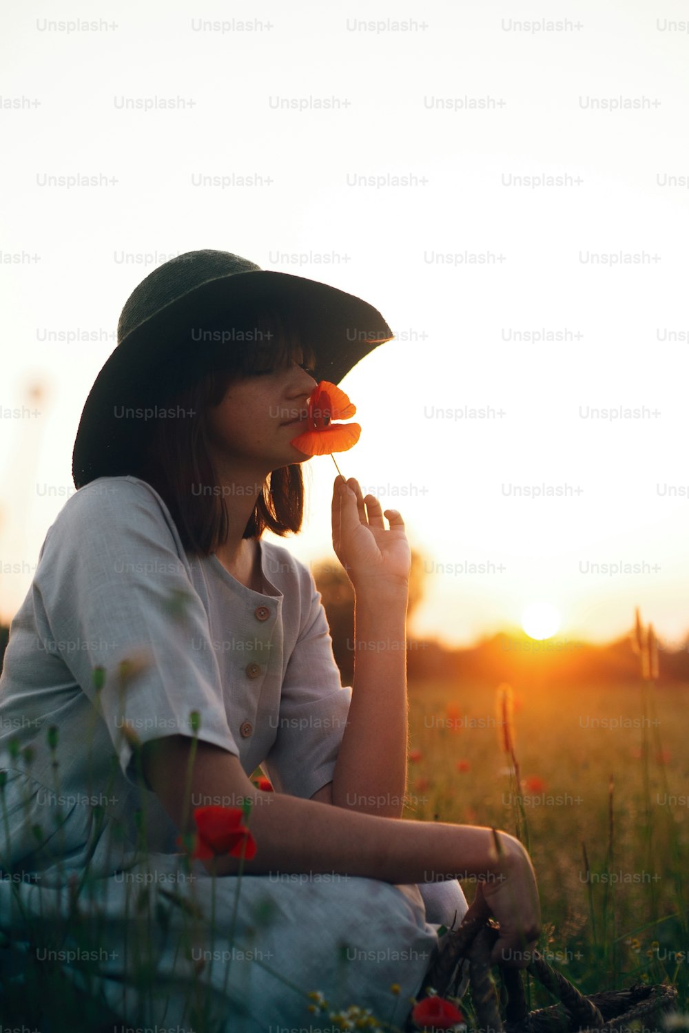 Silueta de una chica elegante en vestido de lino sosteniendo una flor de amapola en el prado a la luz del sol con flores en una cesta de paja rústica. Mujer boho en sombrero relajándose en el campo de la puesta del sol del verano. Atmosférico