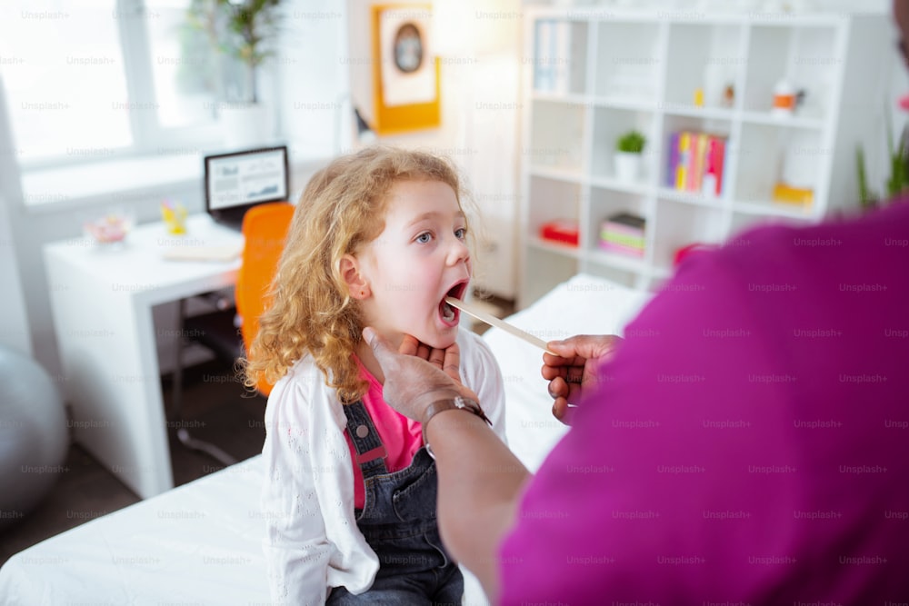 Opening mouth. Cute curly girl having some sore throat opening mouth wide while visiting doctor