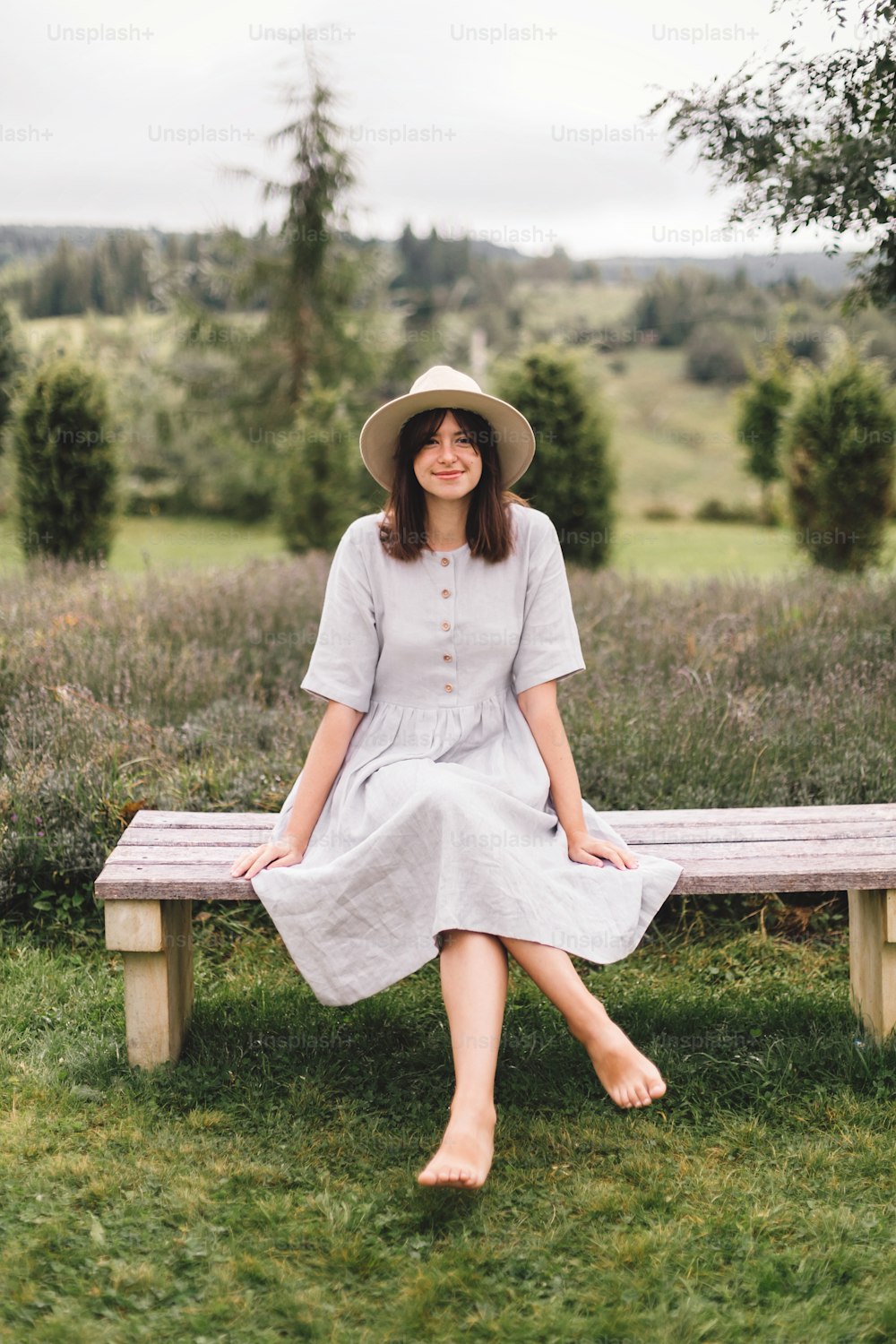 Stylish hipster girl in linen dress and hat sitting on bench at lavender field and relaxing in the morning. Happy bohemian woman enjoying vacation in mountains. Atmospheric rustic moment