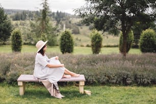 リネンのドレスと帽子をかぶったスタイリッシュなヒップスターの女の子がラベンダー畑のベンチに座って朝にリラックスしています。山で休暇を楽しむ幸せな自由奔放な女性。雰囲気のある素朴な瞬間