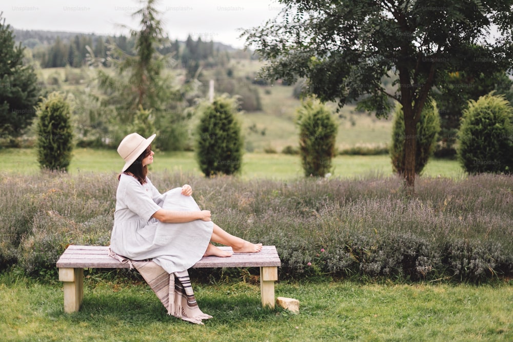 リネンのドレスと帽子をかぶったスタイリッシュなヒップスターの女の子がラベンダー畑のベンチに座って朝にリラックスしています。山で休暇を楽しむ幸せな自由奔放な女性。雰囲気のある素朴な瞬間