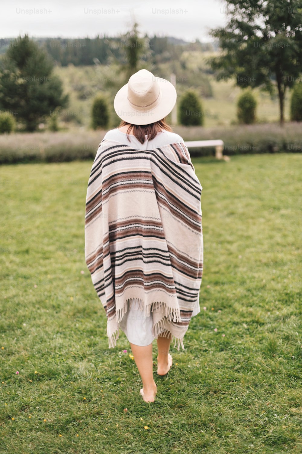 Stylish hipster girl in poncho, linen dress and hat walking at lavender field and relaxing in the morning. Bohemian woman enjoying vacation in mountains. Atmospheric rustic moment. Back view