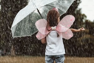 Little girl wearing fairy costume catching water drops with hands outside. She is standing with turned back and holding umbrella with content