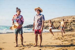 Group of friends have fun together at the beach - men standing and girls behind running to them to play - summer vacation beach concept - millennial people with coloured clothes