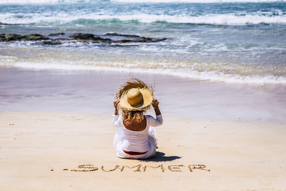 人々との夏休みの休日のコンセプト - 白いドレスの観光客の女性は、海から青い波を見て、帽子を保持して砂浜に座って - 夏は砂の上に書く
